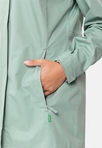 Giacca impermeabile verde chiaro con finitura textured, tasche laterali e polsini regolabili. Piccola etichetta verde visibile.