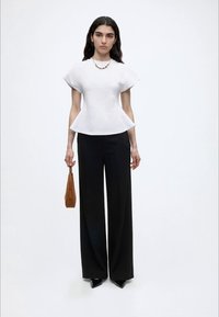 Γυναίκα με μακριά σκούρα μαλλιά που φοράει λευκό εφαρμοστό τοπ, μαύρο παντελόνι με φαρδιά πόδια, μαύρα μυτερά παπούτσια και κρατά καφέ τσάντα.