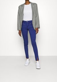 Blå tajta jeans med sidslitsar, kombinerat med en grå rutig blazer och en vit t-shirt. Modell som bär vita sneakers.