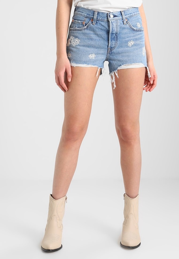 Femme portant un short en denim bleu clair avec une broderie florale blanche et des bottines beiges, debout devant un fond uni.
