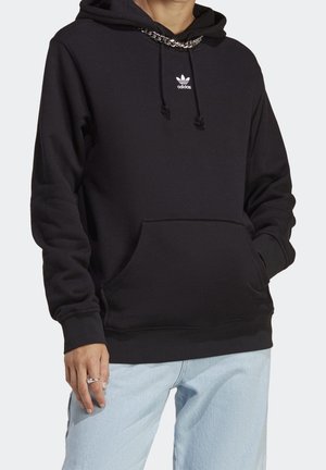 Personne portant un sweat à capuche noir Adidas avec une poche avant et des cordons, associé à un jean bleu clair et un collier chaîne en argent.