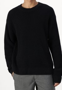 Maglione nero lavorato a maglia con un motivo testurizzato, scollo rotondo e polsini e orlo a coste, abbinato a pantaloni grigi.