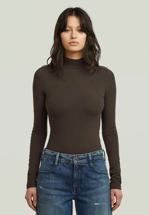 Junge Frau mit langen dunklen Haaren, die einen eng anliegenden braunen Rollkragenpullover und blaue Jeans trägt, steht vor einem schlichten hellen Hintergrund.