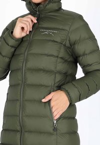 Grüne Steppjacke mit durchgehendem Reißverschluss und Seitentaschen. Ausgestattet mit hohem Kragen und horizontalen Steppnähten zur Isolierung. Logo auf der Brust.