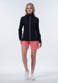 Veste noire zippée avec un col haut, en tissu texturé, et zippers argentés, associée à un short rose clair et des baskets blanches.
