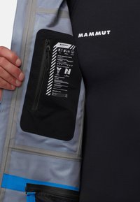 Blue and gray jacket fabric with a black zippered pocket displaying safety information. Texture is smooth with visible branding and hardware.