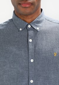 Mężczyzna w niebieskiej koszuli zapinanej na guziki z kołnierzykiem typu button-down oraz małym, żółtym haftowanym logo na piersi.