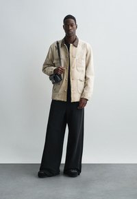 Giacca di jeans beige, pantaloni neri larghi e sneakers scure. La giacca presenta tasche frontali e un colletto a contrasto. Borsa a tracolla nera minimalista.