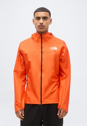Homme portant une veste North Face orange vif avec capuche et fermeture éclair noire, debout devant un fond clair uni.