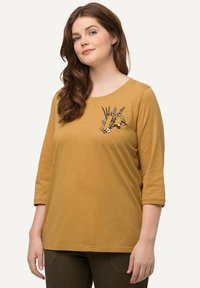 Camisa mostaza amarilla de manga 3/4 con cuello redondo, que presenta un acento bordado floral en el lado izquierdo. Textura de tela suave.