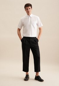Korte mouwen witte overhemd met een borstzak, gecombineerd met zwarte geplooide broek en zwarte veterschoenen, tegen een neutrale achtergrond.