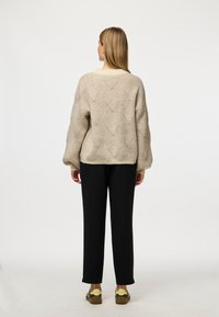 Beige strikkegenser med et strukturert diamantmønster, rund hals og senkede skuldre, sammen med svarte rette bukser.