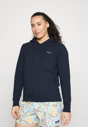 Sudadera con cremallera en color navy, con cordones, bolsillos frontales y un pequeño logo en el lado izquierdo del pecho. Combinada con pantalones cortos estampados con colores pastel.