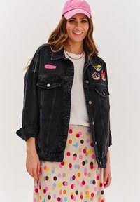 Czarna denimowa kurtka z zapięciem na guziki i haftowanymi łatami postaci na klatce piersiowej, noszona na białej bluzce i spódnicy w kropki. Różowa czapka.