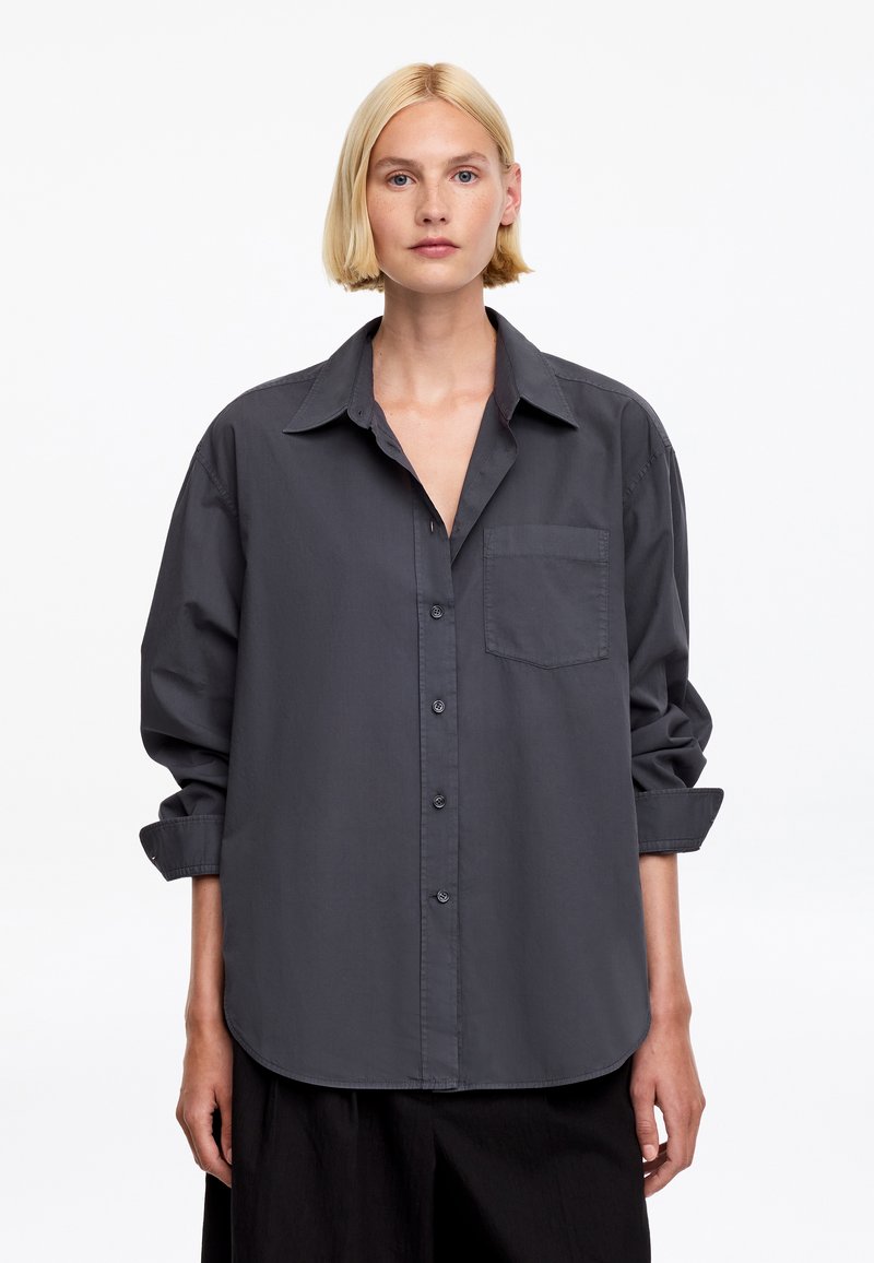 Femme blonde portant une chemise ample gris foncé boutonnée, manches retroussées et une poche avant, debout devant un fond blanc uni.