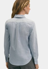 Světle modrá, dlouhá košile s knoflíky vyrobená z hladkého materiálu. Má zadní sklady, kulatý lem a manžety s knoflíkovým zapínáním.