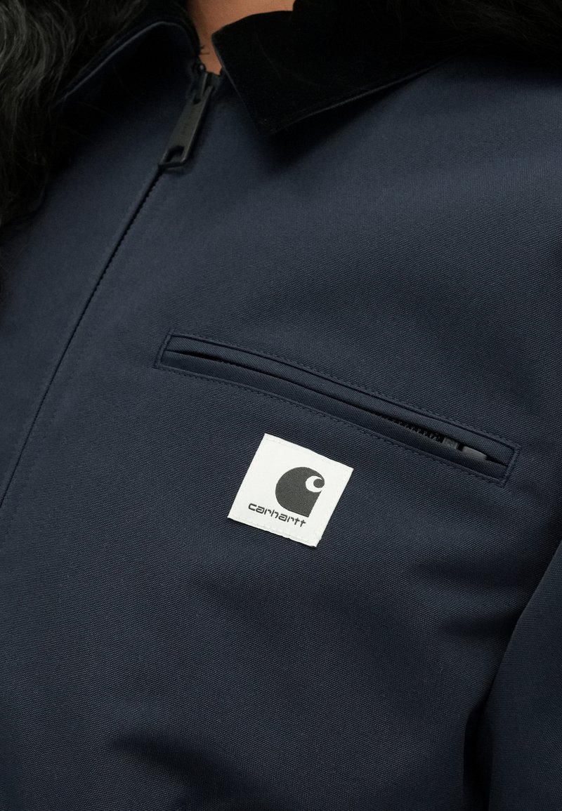 Marineblå jakke med lynlås, der har en sort fløjlskrave, en brystlomme og et hvidt kvadratisk logo på venstre side.