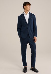Abito blu con giacca su misura e pantaloni slim-fit, abbinato a una camicia bianca. La giacca presenta due tasche frontali, un design elegante e una texture sottile.