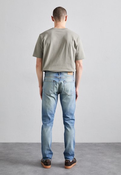 Gray cotton t-shirt, blue faded denim jeans, and brown footwear. The jeans have a round label on the back waistband and classic rear pockets.