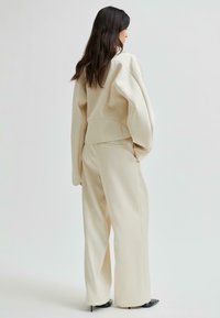 Kvinna med långt mörkt hår som bär en krämfärgad oversized jacka och vida byxor, stående och vänd åt vänster mot en enfärgad bakgrund.