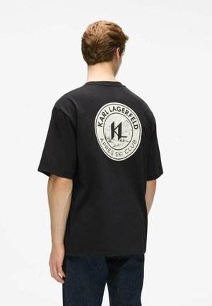 Jeune homme aux cheveux courts et bruns portant un t-shirt noir Karl Lagerfeld Apres Ski Club, vu de dos, se tenant devant un fond uni.