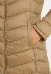 Beige pufferjacka med kviltad design, med dragkedja och sidoficka. Materialet är slätt och något glansigt med en vadderad textur.