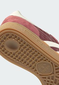 Tenisica s reljefnim gornjim dijelom u boji tamnocrvene i krem, s gumenim potplatom boje gume. Bijele pruge naglašavaju bočne strane, a na potplatu je vidljiv logo.