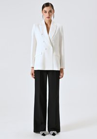 Dubbelzijdige witte blazer met notched revers en twee voorkeurszakken, gecombineerd met hoge zwarte broek en versierde zwarte hakken.