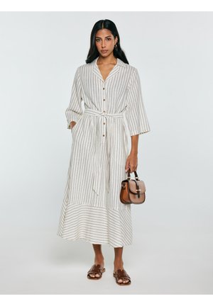 Kobieta w białej, długorękawowej, paskowanej sukience midi z guzikami i paskiem, trzymająca brązową torebkę, stoi w brązowych sandałach.