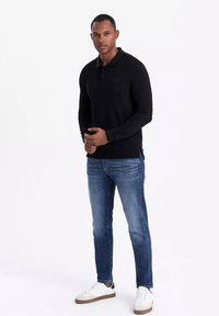 Svart långärmad polo tröja med knäppning, texturerad tyg, sidofickor, matchad med blå jeans och vita sneakers med bruna detaljer.