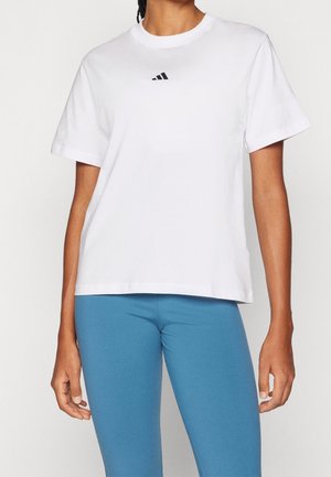 Kobieta w luźnym białym T-shircie Adidas zestawionym z dopasowanymi niebieskimi legginsami, stojąca na prostym jasnym tle.