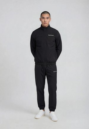 Joven de pie con las manos detrás de la espalda, vistiendo un chándal negro Sergio Tacchini y zapatillas blancas sobre un fondo liso gris claro.