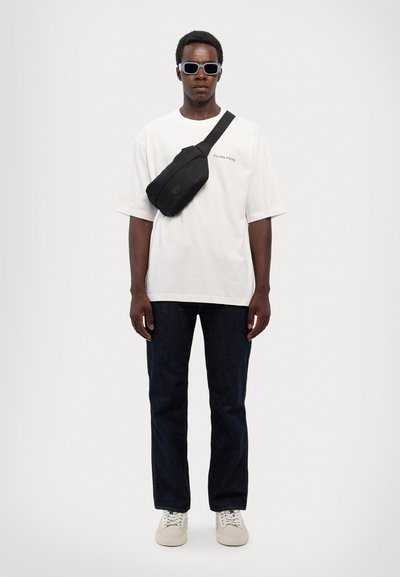 White oversized t-shirt with "Falling Pieces" text, black crossbody bag, dark denim jeans, and beige sneakers; model wearing white sunglasses.