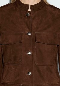 Veste en daim marron avec col mandarin, dotée de deux poches avant, de boutons en métal et de détails cousus. Texture lisse.