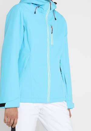 Veste à capuche bleu clair avec fermeture éclair devant, fermeture éclair sur la poche poitrine et sangles ajustables aux poignets, portée avec un pantalon blanc.