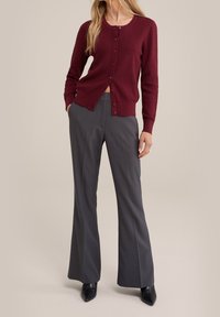 Vinröd stickad kofta med rund halsringning och knäppning framtill, kombinerad med grå utsvängda byxor och svarta spetsiga klackskor.