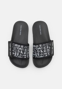Černé pantofle s texturovanou stélkou a širokým popruhem zdobeným bílým značením Calvin Klein Jeans a různými vzory loga.
