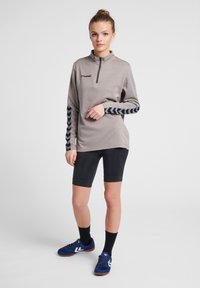 Maglia grigia a maniche lunghe con mezza zip, maniche con motivo a zigzag nero, shorts neri e scarpe da ginnastica blu con dettagli bianchi.