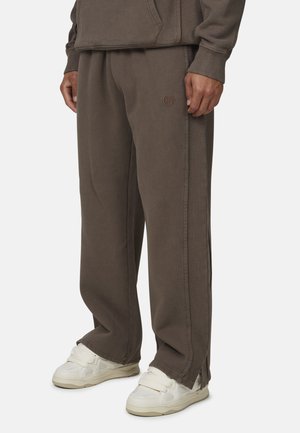 Pantaloni della tuta marroni realizzati in tessuto morbido, con una linea dritta, tasche laterali e un logo discreto sul lato sinistro. Abbinati a sneakers bianche.