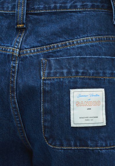 Πίσω τσέπη από σκουρόχρωμο τζιν με πορτοκαλί ραφές και λευκή ετικέτα που γράφει «Summer Vibration with SANDRO 1984 Boulevard Haussmann Paris VIII.»