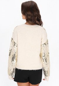 Krem színű kötött pulóver, az ujjakon rojtos arany és fekete flitterekkel díszítve. Puha tapintású, kerek nyakkivágású és laza fazonú.