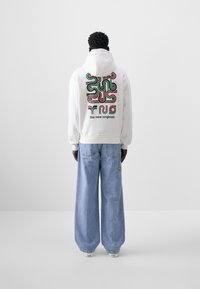 Hvit hettegenser med stort, fargerikt grafisk design på ryggen, sammen med lyseblå vide jeans. Tekst "de nye originalene" inkludert.