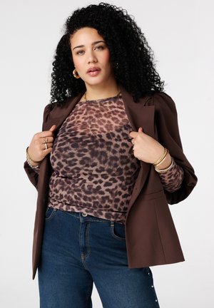 Femme aux cheveux bouclés portant un haut imprimé léopard, un blazer marron, un jean bleu et des bijoux en or, tenant les revers du blazer contre un fond uni.