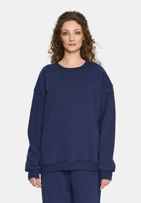 Kobieta z kręconymi włosami, ubrana w luźną granatową bluzę i pasujące spodnie, stoi na tle jasnej białej ściany.