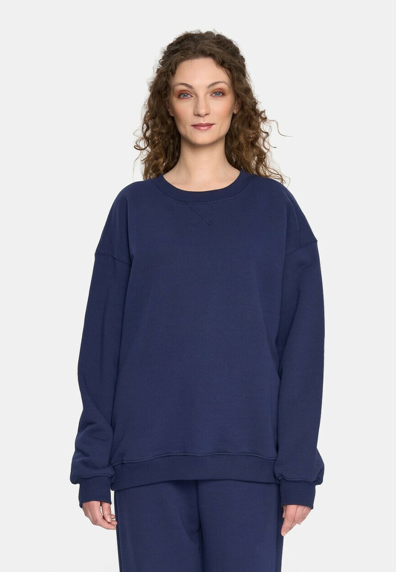 Kobieta z kręconymi włosami, ubrana w luźną granatową bluzę i pasujące spodnie, stoi na tle jasnej białej ściany.