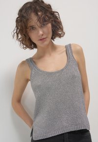 Femme aux cheveux bouclés portant un débardeur en tricot argenté sans manches et un pantalon sombre, se tenant devant un fond clair uni.