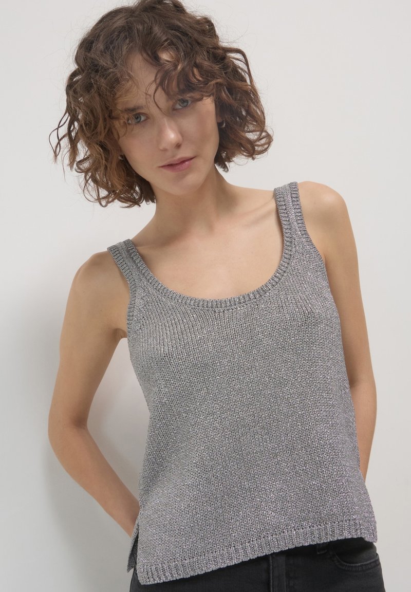 Femme aux cheveux bouclés portant un débardeur en tricot argenté sans manches et un pantalon sombre, se tenant devant un fond clair uni.