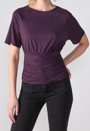 Femme portant un haut violet ajusté à manches courtes avec taille froncée, associé à un jean noir, la main posée sur la hanche.