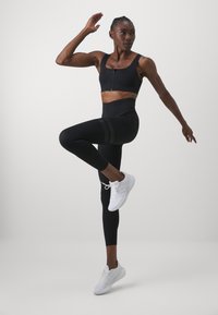 Melna sporta krūšturis ar priekšējo rāvējslēdzēju, augsto vidukli melni legingi ar teksturētām svītrām un baltas sporta kurpes.