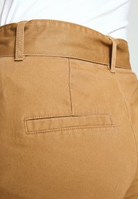 Detailní záběr na béžovou kapsu kalhot se švy a poutek na opasek, nošenou s bílou košilí zasunutou dovnitř.